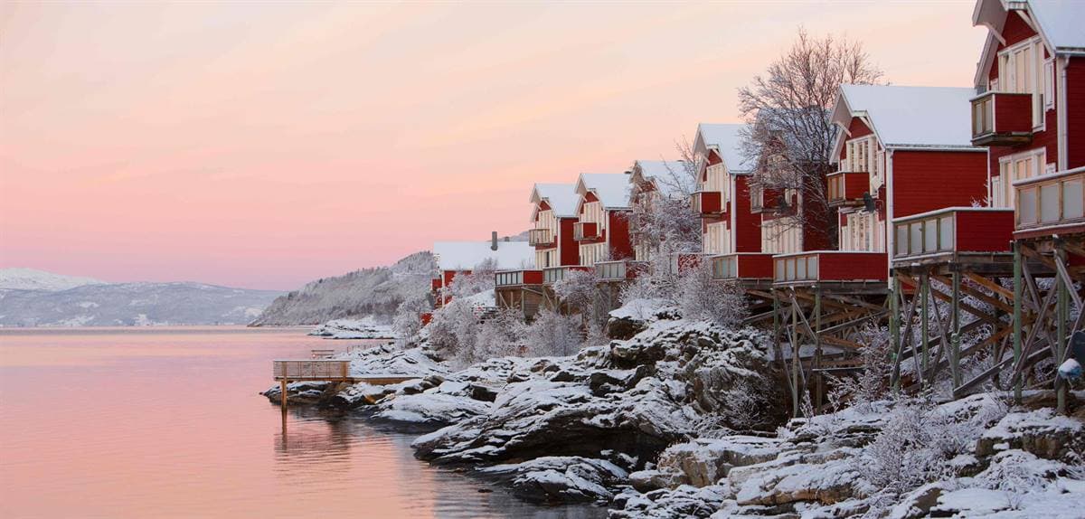 Røde rorbuer på påler på de snødekte svabergene i rosa vintersolnedgang