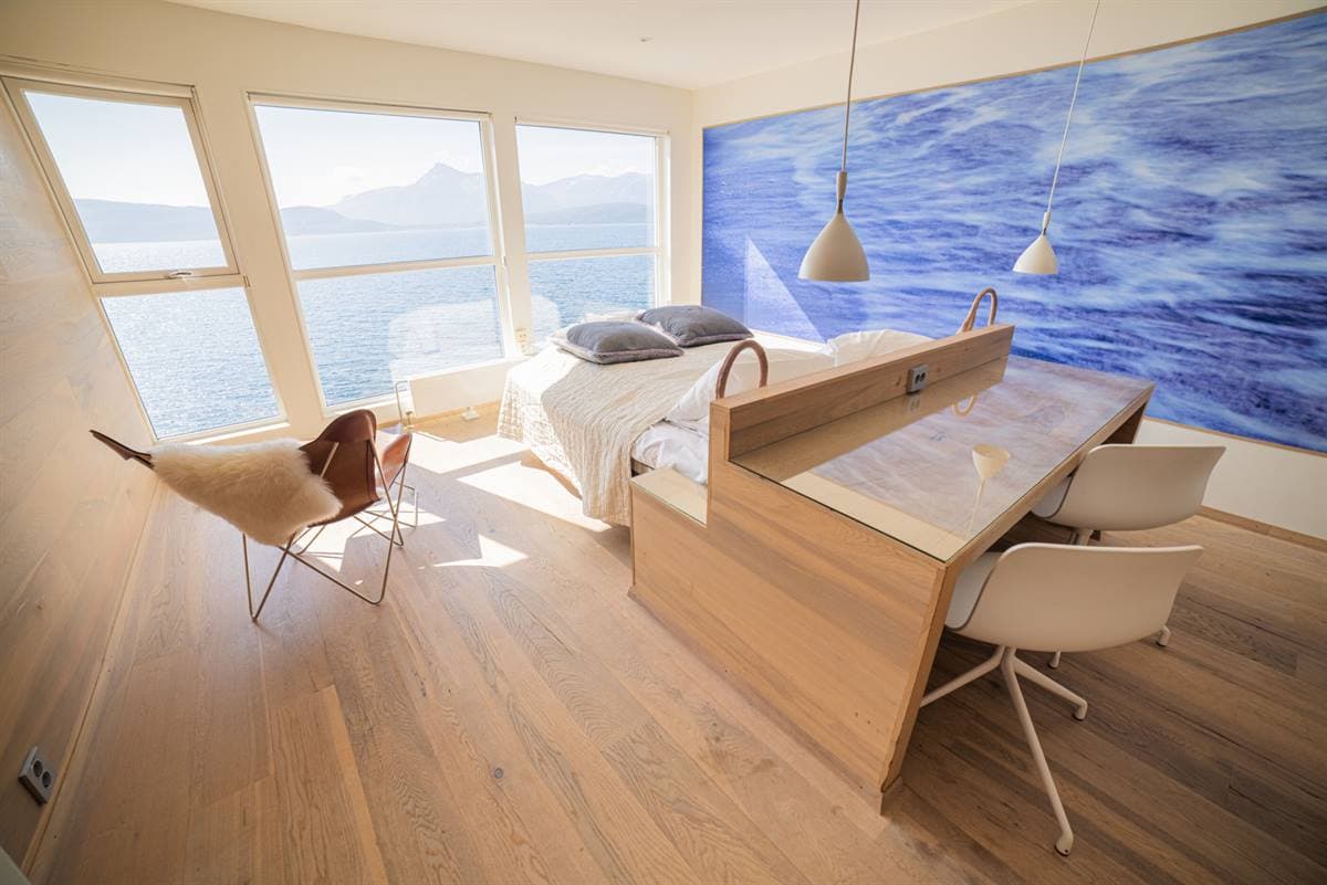 Skrivebord og seng under et bølgemaleri på veggen, med vinduer mot fjorden