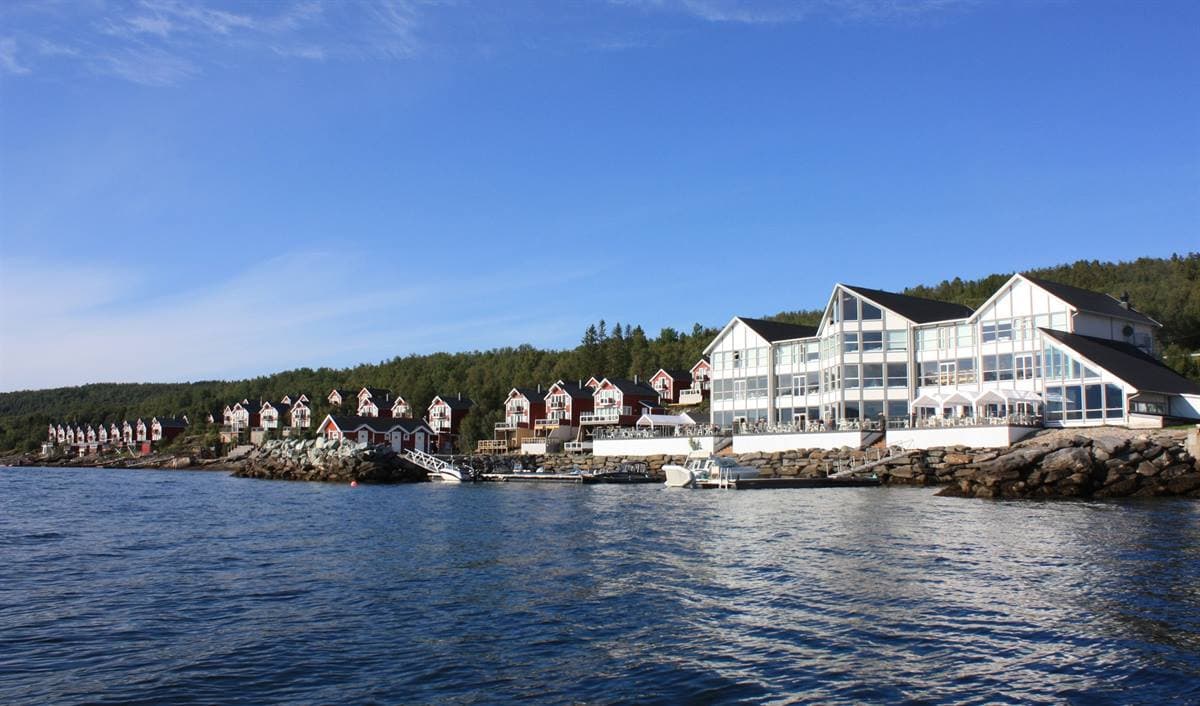 Malangen Resort sett fra fjorden en sommerdag, med det hvite hovedbygget og røde rorbuer langs stranden